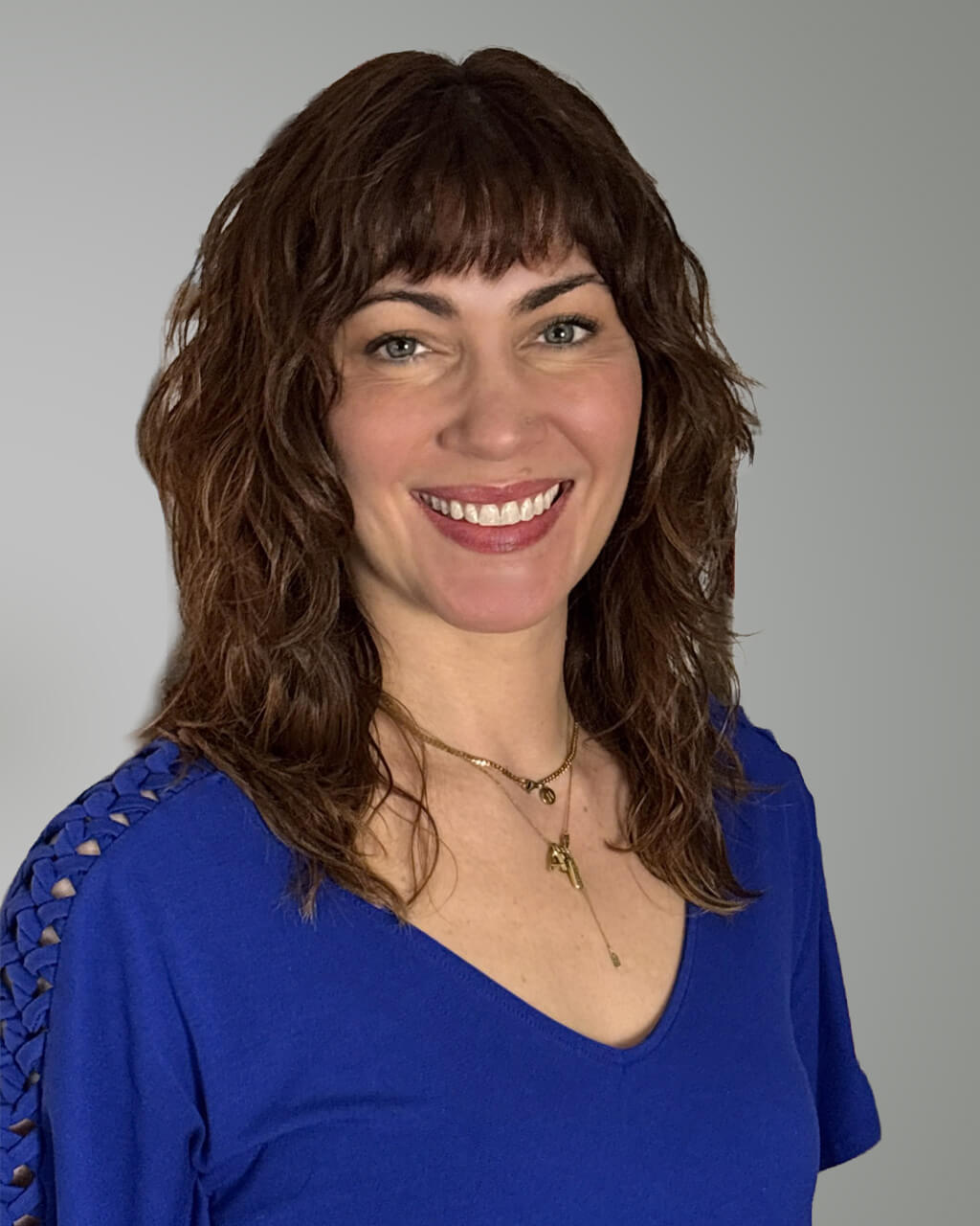 Jennifer Bonino Sobol, a woman with wavy brown hair, wears a blue top and gold necklaces as she smiles at the camera against a neutral gray background.