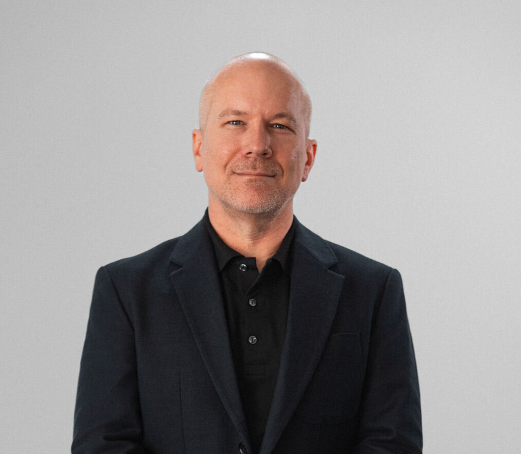 Aiken Benjamin, a middle-aged man with a shaved head and trimmed beard, wearing a black blazer and black shirt, stands against a plain light grey background.