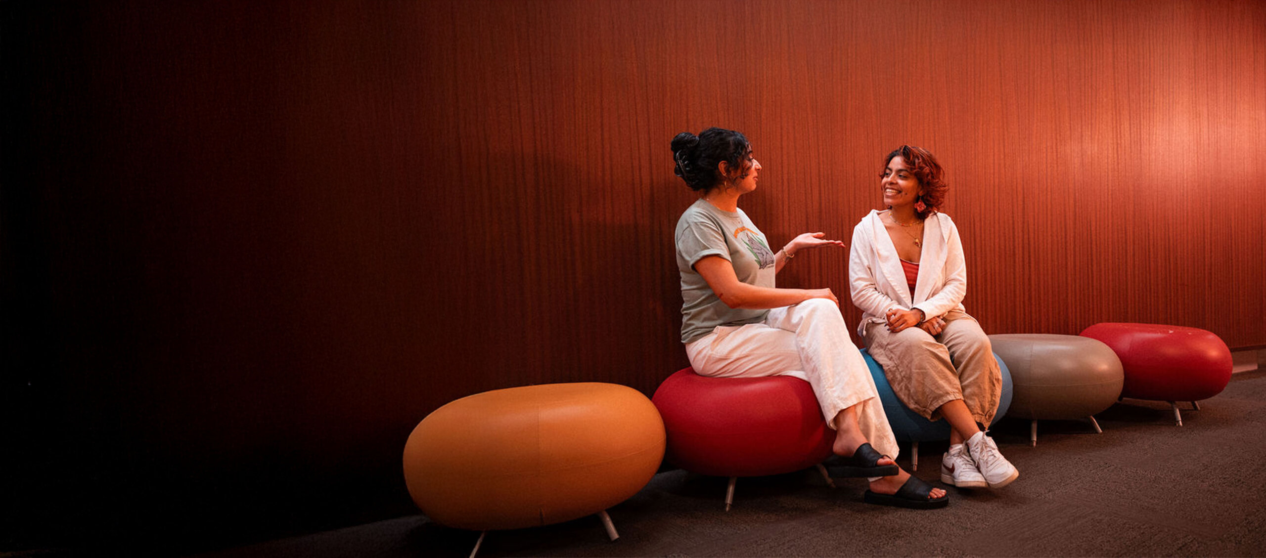 Deux étudiants de l'Université Adler assis sur des tabourets ronds et colorés en train de converser.
