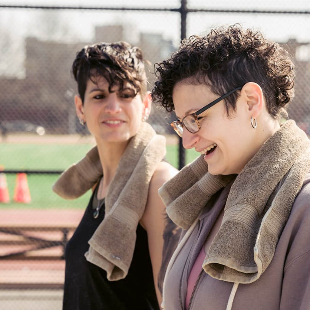 Two people with short curly hair walk outdoors with towels around their necks, smiling and talking about sport on a sunny day near a sports field with a chain-link fence in the background.