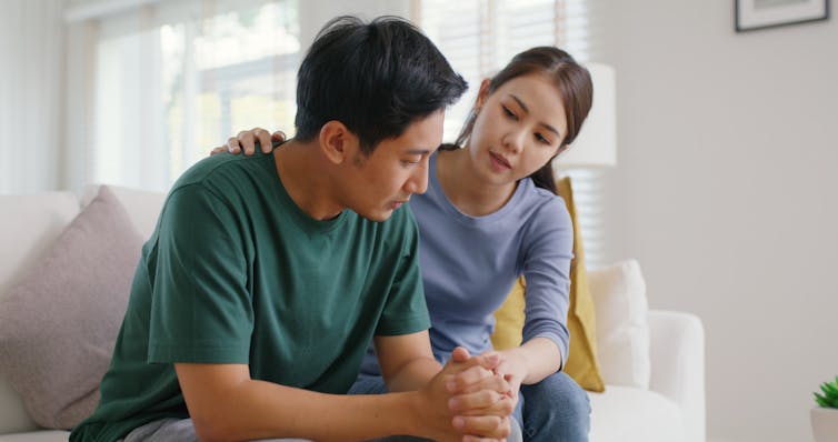 A woman sits beside a man on a couch, gently placing her hand on his shoulder as he looks down with a concerned expression.