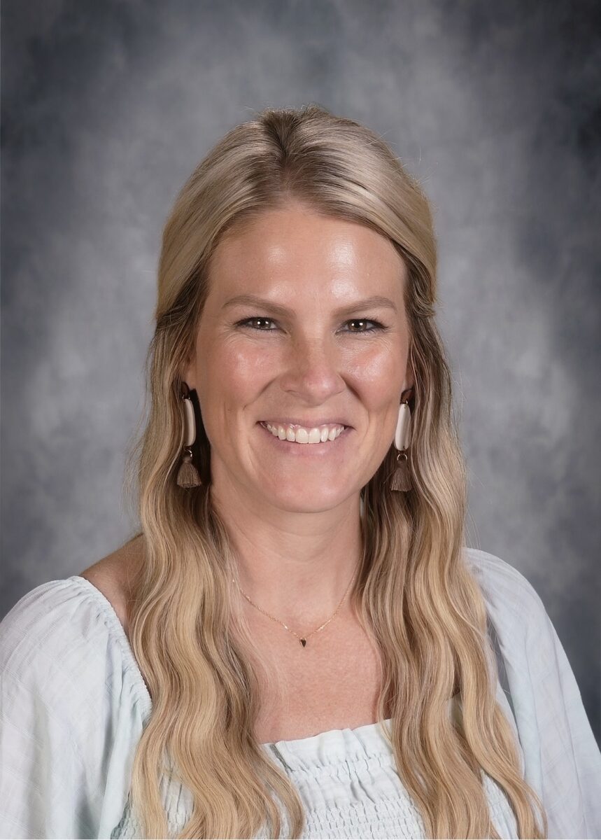 A woman with long blonde hair wearing a light-colored top and earrings smiles in front of a gray mottled background, radiating warmth perfect for sharing stories for children.