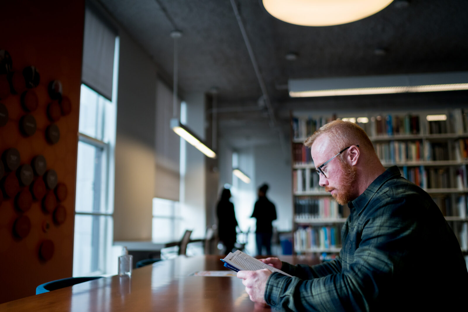 A man with glasses sits at a table in a library, reading a small book—a quiet moment of student life, with bookshelves and two people blurred in the background.
