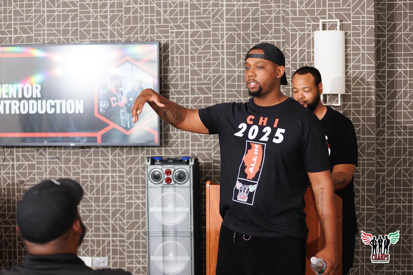A man wearing a "CHI 2025" shirt, an Adler alum, speaks about how mentorship transforms lives to a seated audience, while another man stands behind him at a podium. A screen reads "Mentor Introduction.
