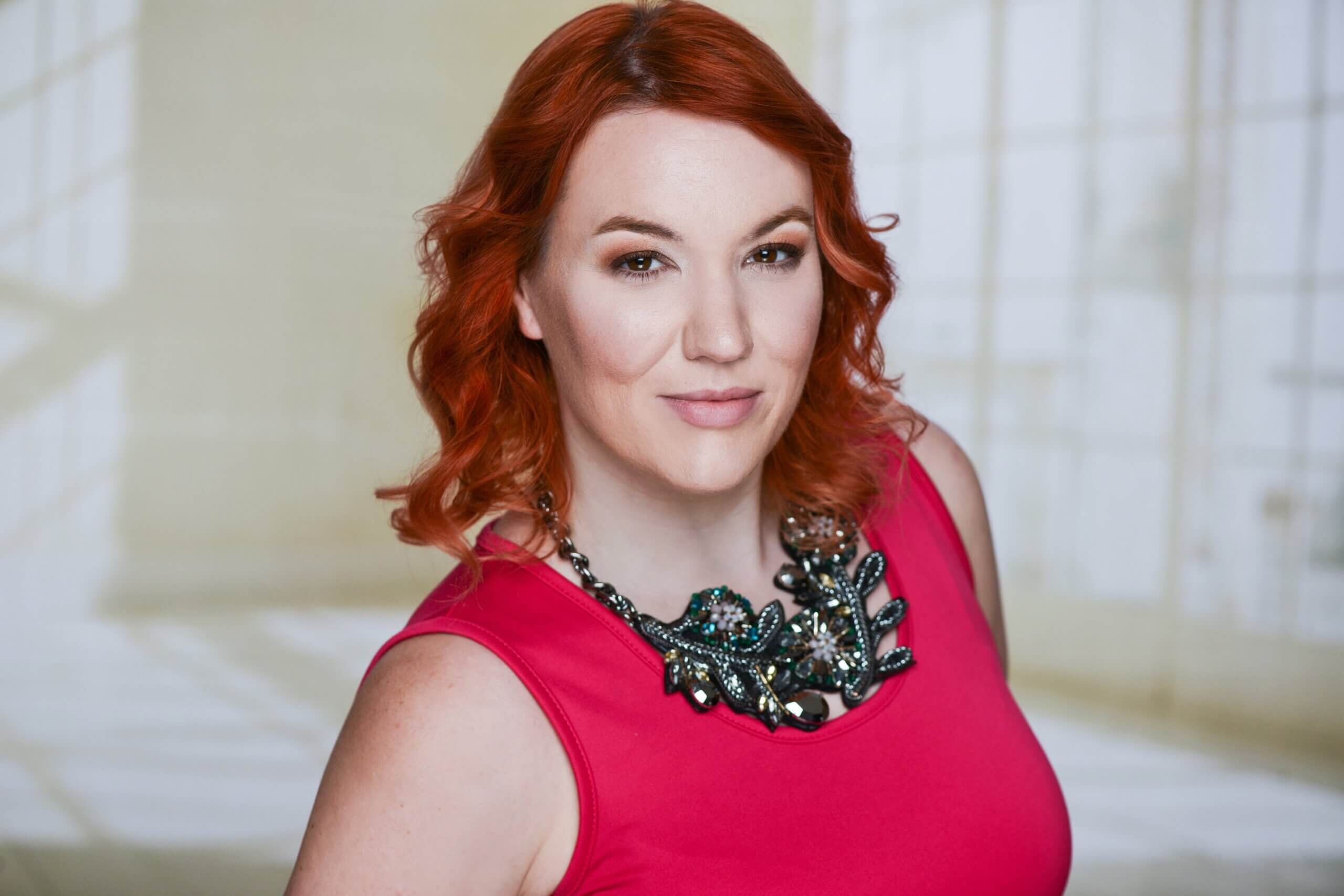 A woman with wavy red hair wearing a pink sleeveless top and a large statement necklace stands in front of a softly lit background.