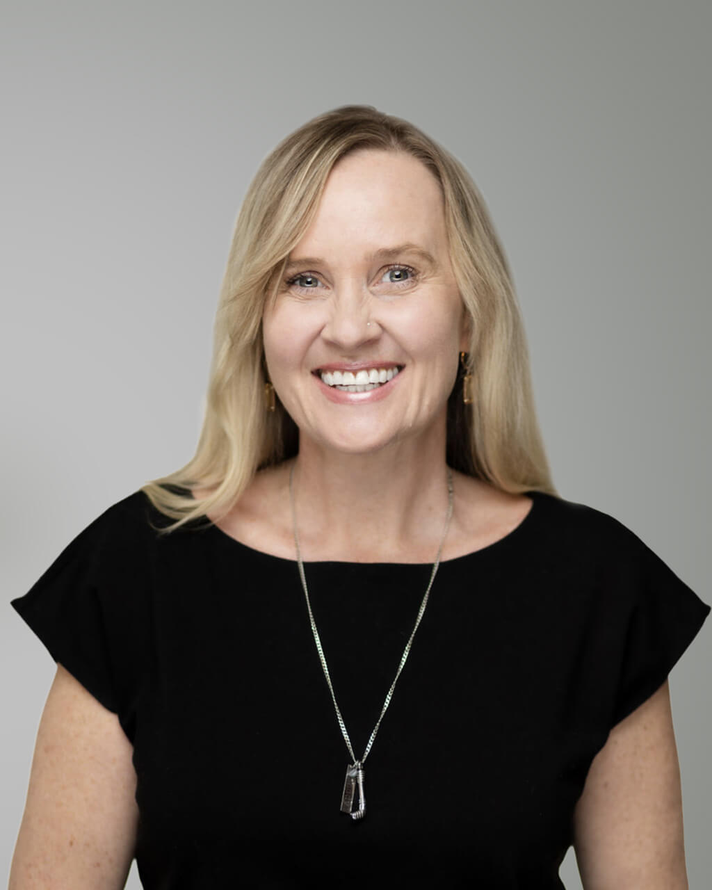 Erica Bearss, a woman with long blonde hair, wears a black top and necklace, smiling at the camera against a plain light gray background.