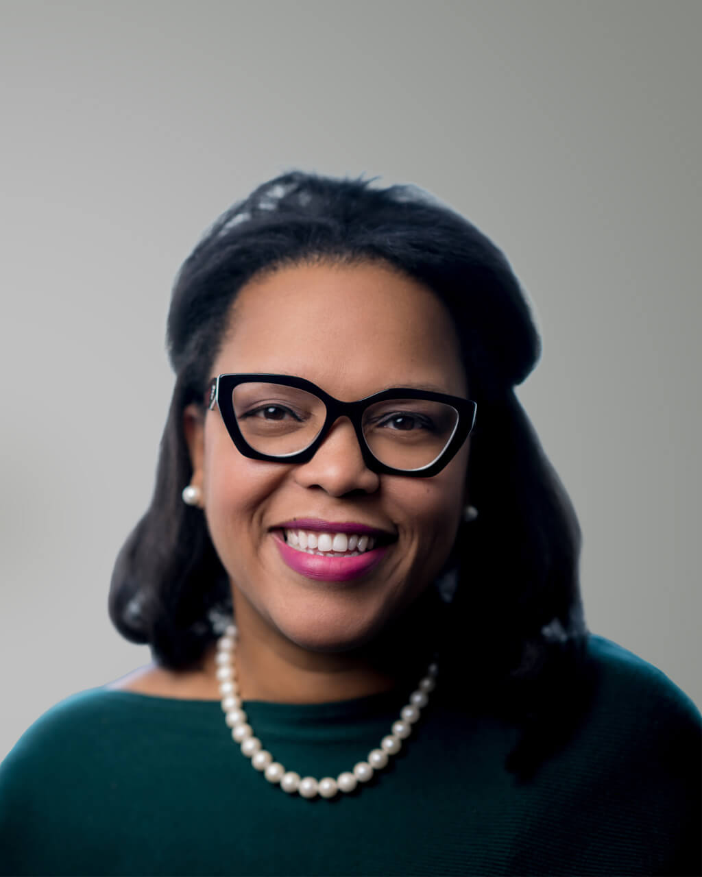 A woman with dark hair, wearing black glasses, a pearl necklace, and a dark top, smiles at the camera against a plain background—introducing Kesha S. Burch.