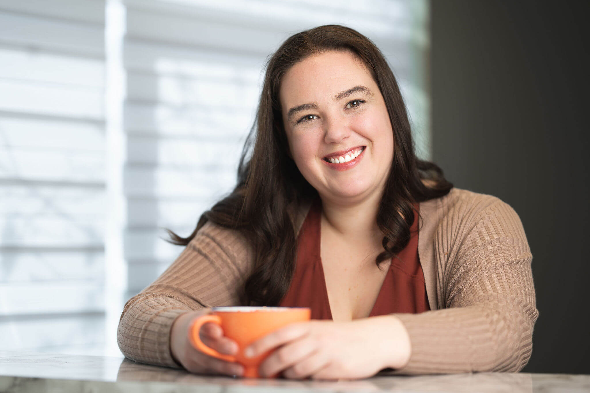 A woman with long brown hair, wearing a brown cardigan, sits at a table holding an orange mug and smiling at the camera, her warmth reminiscent of an Aurora on a chilly Arctic evening.
