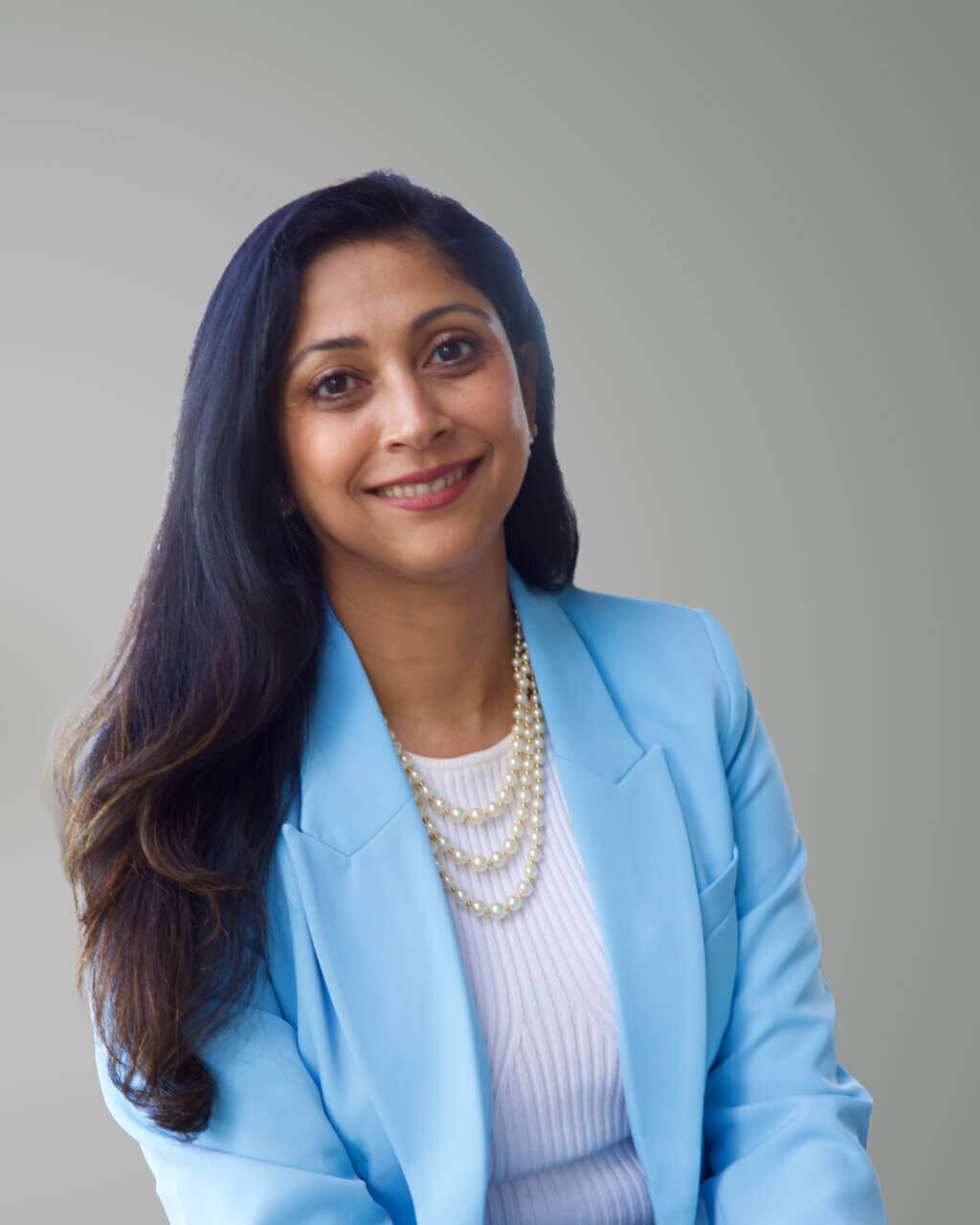 Une femme aux longs cheveux bruns, vêtue d'un blazer bleu clair, d'un haut blanc et d'un collier de perles à plusieurs rangs, sourit avec assurance devant un fond gris clair uni — alliant style et assurance dans le monde du travail d'aujourd'hui, où l'IA impose un rythme effréné.